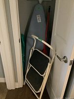 Photo showing ironing board with gray cover, two-step stepstool leaning against it, with dustpan and broom in the background.