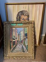 Two framed artworks leaning against the wall. Portrait of woman is in a gold frame, cityscape with ornate frame is loose and leaning in front.