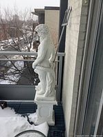 Front view of full-length concrete Poseidon statue on square pedestal, displayed on balcony over snow-covered floor.