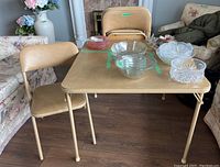 Tan folding card table and one folding chair shown from side angle with items on table.
