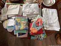 Wide shot showing multiple folded linens on table including white napkins, lace-edged cloths, colorful tea towels, and oven mitts