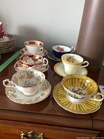 Six tea cups and saucers arranged on wood surface showing variety of floral and gold design patterns