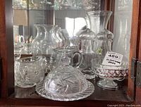 Clear glassware displayed in a cabinet, including butter dish, creamers, salt and pepper shakers, decanters, and vases.