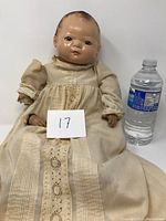 Front view of the antique composite doll wearing a cream-colored dress, seated next to a water bottle for scale. The doll has a composite head and hands with visible facial details and molded hair.
