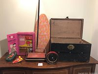 Photo showing the open black vintage child’s trunk, Bissell Little Queen iron, child’s floral ironing board, boxed School Closet toy set, and several small toys on a wooden surface.