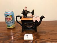 Front and side view showing the black ceramic sewing machine shaped tea pot with pink cloth and small package on top, next to a soda can for size comparison.