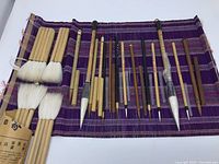 Full set of 15 brushes on woven purple cloth, showing 4 large Yasutomo hake brushes with thick white hair and 11 smaller bamboo handle writing brushes all with lids.