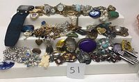 Wide view of assorted jewelry on white riser including watches, brooches, necklaces, and pins in various colors and materials