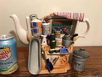Full side profile of the large cream-colored Paul Cardew teapot decorated with miniature colorful garden tools and accessories, placed next to a soda can for scale.