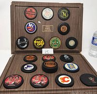 Photo showing two wooden display holders opened to reveal 19 vintage hockey pucks with various team logos and commemorative markings.