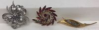 Photo showing three brooches in a row, from left to right: large silver-tone flower brooch, red garnet swirl brooch, and gold-tone pod brooch with green and blue stones.