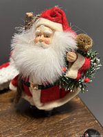 Close-up of large Santa Claus figurine showing detailed face, white fluffy beard, and red plush suit holding sack with greenery and pine cone.