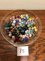 Top-down view of approximately 85 marbles arranged in a glass dish (dish not included in lot) showing varied colors and sizes.
