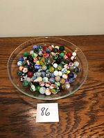 Marbles in a clear, shallow dish on a wood surface, showing variety in color, size, and pattern.
