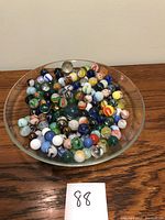 Top-down view of assorted glass marbles of various colors and sizes in a glass dish on wood surface. Number 88 card present indicating lot number.
