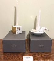 Two signed Rosenthal Studio Line boxes with one square and one round white porcelain candle holders topped with white candles. The square holder shows a single angel illustration in gold and black, the other a circular holder with multiple angel faces and a wing-shaped handle.