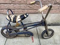 Side view of the small vintage child's bike showing blue metal frame, worn padded seat with torn cover, rubber wheels, and handlebar grips.