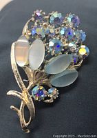 Front view of vintage flower bunch brooch showing blue opalescent oval stones and purple iridescent rhinestones on silver-tone metal base.