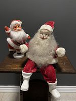 Photo of both vintage Santa figurines positioned on a wooden ledge; one Santa sitting with porcelain head and cloth suit, the other standing on white plastic base.