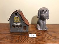 Front view of the barn-shaped vintage tobacco advertising coin bank and the gray elephant rubber coin bank on a wooden surface.