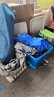 Photo showing blue tent bags, blue plastic tote with tent fabric, beige foldable garden kneeler, and green garden tool boxes partially visible.
