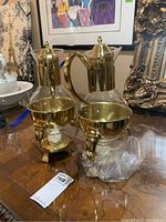 Two brass and glass coffee carafes with candle warmers on a wooden surface, showing original tags and packaging material.