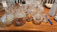 Full lot of mixed clear glassware on wood table, showing covered boxes, toothpick holder, decanter, finial, and bowl