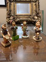 Four Goebel figurines Little Helper, Lost Stocking, Little Gardener, Boy with Knapsack displayed on a glass table with ornate gold picture frame in background.