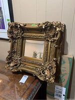 Front angled view showing detailed gold painted frame with carved scrolls and floral motifs around beveled square mirror