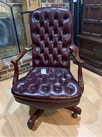 Front view of burgundy leather-like tufted office chair showing seat cracking, wood arms, nailhead trim accents, and caster wheels.