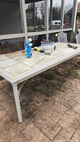 View of rectangular patio table with tiled top and metal legs, outdoor setting, showing tile surface and metal frame.