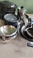 Overview of entire cookware set showing enamel stockpots, frying pans, wok, stainless steel steamer insert, and lids arranged on floor.