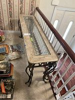 Full view of the metal and glass sofa table next to staircase railing, showing the entire length and top beveled glass insert surrounded by a distressed finish surface.