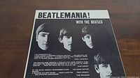Front cover of the album 'Beatlemania! With The Beatles' showing black and white portraits of The Beatles members and text reviews.