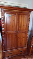 Front view of closed wooden armoire showing two paneled doors and bottom drawer with metal handles. Carved detail on sides and top molding visible.