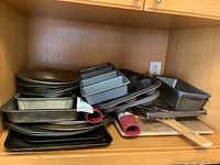 Photo of assorted metal bakeware including loaf pans, cookie sheets, muffin tins, pie pans, pizza pans, silicone mats and cooling racks inside wooden cabinet.