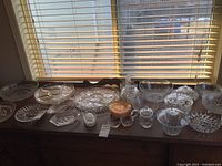 Photo of various clear glass, crystal lidded jars, serving platters, bowls, small basket, and carnival glass saucers arranged on a wooden surface in front of window blinds