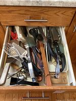 Opened drawer showing a variety of kitchen utensils including whisks, spatulas, tongs, and knives in a cluttered arrangement.