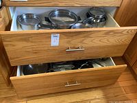 Two wooden drawers filled with assorted stainless steel pots, pans, and lids with some showing wear and scratches.