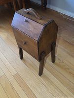 Full view of solid wood sewing stand showing angled drop-lid with knob, carrying handle, and four legs on hardwood floor.