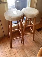 Pair of stools with woven fabric seats and round wooden frames, placed near a window on a hardwood floor.