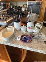 Photo of various kitchen utensils and bowls arranged on a kitchen counter, showing black plastic utensils in ceramic holders, metal ladle and strainer, and multiple ceramic items including bowls and resting spoons.
