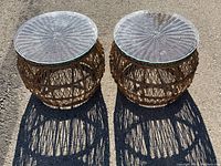 Photo showing two round wicker side tables with glass tops side by side on asphalt surface in sunlight.