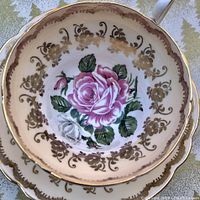 Close up top-down view of tea cup and saucer showing large pink rose center and gold filigree pattern on white porcelain.