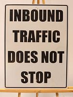 Front of a rectangular enamelled metal sign with bold black lettering reading 'INBOUND TRAFFIC DOES NOT STOP' on a white background with black border, mounted on an easel.