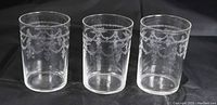 Three vintage clear glasses with fine etched garland designs around the top band, arranged in a row on dark background, showing full set.