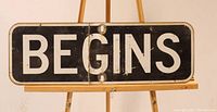 Front of metal sign with white 'BEGINS' text on black enamel background, showing crease and mounting holes.