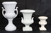 Three white ceramic classical urn flower vases of varying sizes with two side handles, captured together on black background.