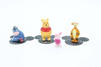 Front view of all four ceramic figures arranged in a row with white background, showing Eeyore, Winnie the Pooh, Piglet, and Tigger; Eeyore, Pooh, and Tigger on black Mickey Mouse base, Piglet without base.