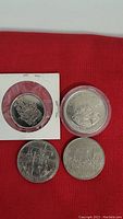 Four silver-toned Canadian coins on red background including a paper held 50 cent coin, a plastic holder $1 coin, and two other $1 coins.